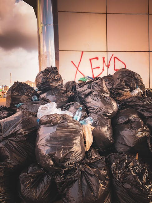 A large pile of black rubbish bags, some partially torn or bulging, stacked against a modern building with a tiled cladding surface. The bags appear full and contain various waste materials, with a few transparent plastic bottles and other litter visible among them. Behind the pile, red spray-painted graffiti spelling 'XEND' is visible on the building's tiles. The scene is outdoors, possibly on a pavement or driveway near an urban setting, with a cloudy sky and distant cityscape visible in the background. The overall impression is of unmanaged waste accumulation, highlighting the importance of private rubbish removal services such as those provided by House Clearance Hounslow, which offer alternative disposal options outside of local authority collection channels. The lighting suggests late afternoon with a slight warm tone, emphasizing the prominence of the rubbish in the foreground and the reflective surface of the building's finish.