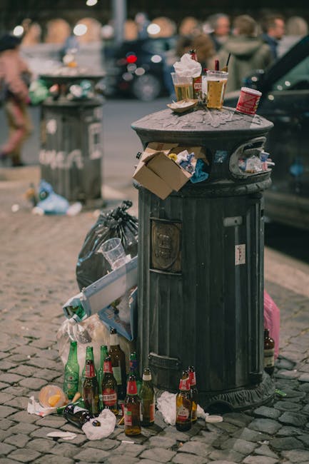 A large accumulation of various waste materials is piled up on a paved sidewalk adjacent to a parking area. The trash includes numerous black, brown, and red rubbish bags, some tightly tied and others torn open, revealing contents such as paper, plastic packaging, and cardboard boxes. Several flattened cardboard boxes and crumpled paper are scattered across the ground. A prominent grey metal recycling bin labeled for mixed paper and card, filled with flattened boxes and loose paper, stands at the center of the heap. Behind the pile, a silver hatchback car is parked, partially obstructed by a metal railing that encloses part of the sidewalk. In the background, a building features storefronts with shopfront signage and large windows, some of which display advertisements. Above, a blue scaffolding structure indicates ongoing maintenance or construction work, and the entire scene is illuminated by natural daylight. This image exemplifies the kind of collection that might require private waste removal services, such as those provided by House Clearance Hounslow, highlighting proper disposal practices for rubbish removal in urban environments.