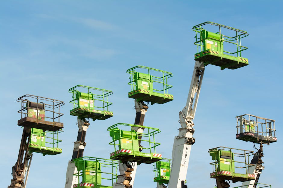A row of multiple aerial work platform boom lifts with articulated arms, positioned upright against a clear blue sky. The lifts have green metal baskets with safety rails, some showing rust or wear on their frames. The platforms are elevated at different heights, with the arms extending upwards and slightly angled, indicating they are stationary or in use for maintenance or access work. The equipment is situated outdoors, likely in a staging area or construction site, with no visible ground or surrounding environment shown. The scene emphasizes the machinery used for height access, relevant to safe rubbish removal or clearance tasks that require reaching difficult or tight spaces, as could be seen in private waste collection contexts. The lighting is natural daylight, highlighting the colors and textures of the lift components and casting minimal shadows, with a focus on the industrial nature of the equipment associated with waste handling services.