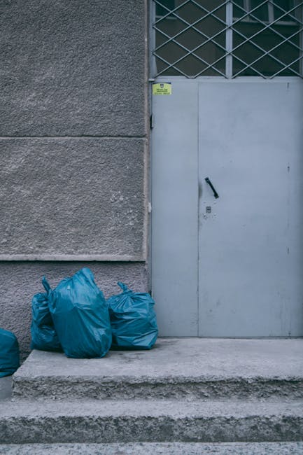 The image shows a set of three blue plastic garbage bags placed on a concrete doorstep in front of a grey metal door, which features a black handle and a small lock. The door is set within a building that has a textured, dark grey exterior wall. To the right of the door, there is a section of window with a diamond-patterned metal grille above it, suggesting an industrial or commercial setting. The scene appears to be outdoors, positioned on a paved area with steps leading up to the door, and the lighting suggests daylight. This visual context aligns with on-site waste management or private rubbish collection services, emphasizing the handling of collected rubbish in an urban environment, and reflects typical components involved in refuse disposal activities that local authorities may not cover. House Clearance Hounslow is involved in waste removal, and the scene exemplifies typical rubbish accumulation for independent disposal or collection efforts in a professional setting.