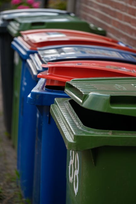 A row of five large household waste bins lined up outside beside a brick wall on a paved surface. The bins are made of durable plastic with textured surfaces and are colored green, blue, and red, each with a matching lid. The green bin at the front has a partially open lid, revealing the interior, while the other bins are closed. The bins appear to be used for recycling and general waste collection, with visible markings and symbols indicating their designated waste types. The environment is an outdoor setting with natural light, and the background includes a brick wall and blurred greenery. This scene illustrates the typical arrangement for rubbish collection areas, which may reflect options for private waste disposal services such as those provided by House Clearance Hounslow, supporting efficient waste management outside residential properties.