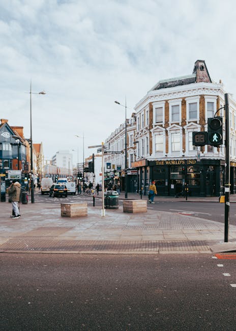 A busy street scene featuring a corner building with a rounded façade, four stories high, painted in white with brown brick accents around the windows, and a ground-floor pub named 'The World's End' with dark signage and large windows. In the foreground, the paved pedestrian area has lightweight wooden planters and street furniture, with a traffic light showing a green pedestrian signal and a crosswalk leading across the intersection. Several pedestrians are walking along the pavement, some waiting at the crossing, while vehicles, including a white van and cars, are visible on the street. Overhead, the sky is partly cloudy, and streetlights and utility poles are positioned along the road, contributing to an urban environment typically associated with a central shopping and social area capable of supporting independent waste collection and rubbish removal services, as provided by companies such as House Clearance Hounslow. The scene captures the vibrancy of an active high street, suitable for matching content about local rubbish clearance and alternative disposal solutions in a city setting.
