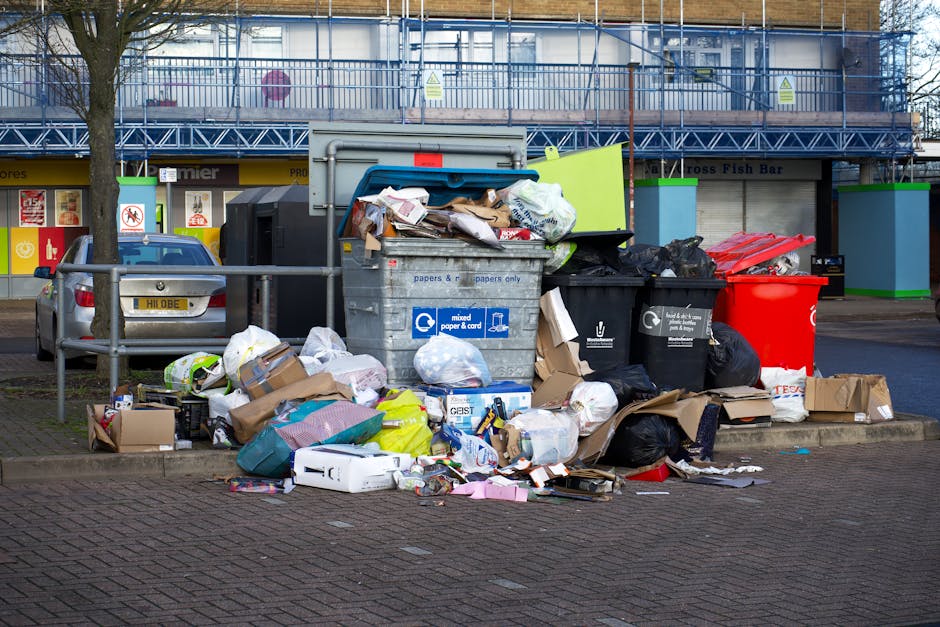 A large pile of mixed waste and rubbish bags is collected in front of a public bin in an urban parking area, with debris including cardboard boxes, paper bags, and plastic packaging spilling onto the paved ground. The waste appears in various materials such as paper, cardboard, plastic, and black garbage bags, some of which are partially open, revealing their contents. Behind the waste, a silver car is parked, and a metal railing separates the rubbish from the vehicle. In the background, there are commercial storefronts with yellow signage and posters, as well as a building under renovation covered with blue safety netting and scaffolding. The overall scene suggests an area where private rubbish collection might take place, as offered by local waste management services like House Clearance Hounslow, with environmental context highlighting the importance of proper rubbish disposal and alternative waste handling methods.