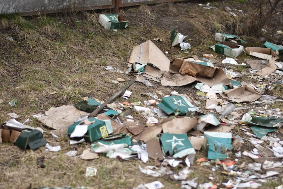 A patch of ground adjacent to a grassy area and a high wooden fence shows scattered rubbish, including torn cardboard boxes, some green with white star logos, and torn paper packaging. The cardboard boxes are crumpled, with some flattened and others partially collapsed, and their surfaces display creases and torn edges. There are also small pieces of paper, plastic wrappers, and debris mixed among the larger items, indicating an untidy and unmanaged waste site. The grass surrounding the litter is patchy with uneven coloration, and the scene appears to be outdoors, possibly alongside a drainage ditch or an unkempt verge. The scattered rubbish suggests a need for alternative waste handling or rubbish removal services such as those provided by House Clearance Hounslow, especially in areas where illegal dumping or accidental waste dispersal occurs. The overall environment appears neglected, emphasizing the importance of proper disposal to maintain cleanliness and environmental standards.
