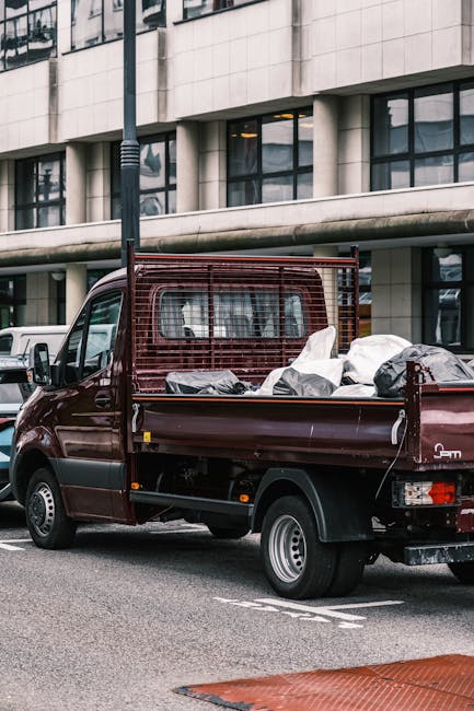 A small, burgundy flatbed truck parked on a city street in front of a modern, multi-storey building with large glass windows and a concrete facade. The truck's cargo area is filled with various black and white plastic bags, some appearing to contain household waste or discarded items. The vehicle features a metal mesh cab guard behind the cab, and its rear lights, wheel arches, and side mirror are visible. Surrounding the truck, there are a few other parked vehicles, including a white van to the left. The roadway surface is dark asphalt with painted parking lines and a red textured pedestrian area in the lower right corner. The environment indicates an urban setting, often associated with private rubbish removal or on-site clearance services, such as those offered by House Clearance Hounslow. The scene captures a typical private waste collection activity aligned with alternative rubbish handling methods outside of local authority waste disposal channels, reflecting the nature of independent rubbish removal in a cityscape context.