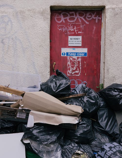 A patch of ground covered with scattered debris and discarded packaging materials, including flattened cardboard boxes with printed logos and text, open and torn, along with crumpled paper and smaller litter fragments. The debris is spread across a grassy area adjacent to a fence, with some of the cardboard boxes damaged and partially flattened, and a metal tool or pole lying among the rubbish. The environment appears outdoor, possibly a neglected or unmanaged site, with uneven terrain and sparse, dull vegetation. The scene highlights a case of improper waste disposal, relating to private rubbish removal services such as those offered by House Clearance Hounslow, and illustrates the need for professional rubbish collection to manage such waste more effectively and avoid environmental contamination.
