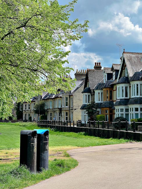 A pair of black waste bins positioned on a paved pathway in a residential area with green grass surrounding them. In the background, there are several Victorian-style terraced houses with bay windows, chimneys, and slate roofs, set behind a low wooden fence. To the left, a large tree with fresh light green leaves extends its branches over the scene, partially shading the pathway. The sky above is partly cloudy with patches of blue visible, creating a bright, natural outdoor setting. The image captures a quiet street scene suitable for private waste collection or local rubbish removal services, with the bins indicating waste disposal points in front of residential properties. House Clearance Hounslow may use such visual cues to illustrate standard rubbish handling and on-site clearance options in suburban environments where independent collection is a common alternative to council services.