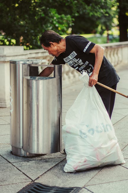 A man with short dark hair, wearing a black T-shirt with white graphic text on the front, is seen disposing of rubbish into a modern, cylindrical stainless steel public trash bin located on a paved sidewalk. He is bent forward, holding a large white plastic sack filled with waste in his left hand, while his right hand is reaching into the bin to deposit more rubbish. The bin is positioned next to a low stone wall with a decorative balustrade, and in the background, there are lush green trees and a grassy area, suggesting an outdoor setting in a park or urban street. The scene is illuminated by natural daylight, creating a clear, bright atmosphere that aligns with private or independent waste disposal services, such as those provided by companies like House Clearance Hounslow, catering to efficient rubbish removal and collection. The image captures an everyday act of waste management, emphasizing cleanliness and proper rubbish disposal in a public space. The surface of the bin has a brushed metal finish, and the gray paving stones beneath the man reflect the natural light while the background foliage provides a contrasting vibrant green backdrop.