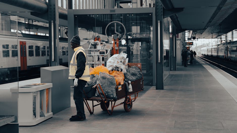 A municipal rubbish collection truck, painted white with some rust visible on its rear loading mechanism, is parked on a narrow cobblestone street in an urban residential area. The truck is in the process of collecting waste, with its rear compartment partially open, revealing the internal hydraulic lifting arms and rusty metal framework. A sanitation worker dressed in blue overalls, an orange high-visibility vest, and a blue cap is positioned at the rear, handling a blue wheelie bin to dispose of waste into the truck’s hopper. The surrounding environment includes multi-story buildings on either side of the street, with weathered facades, some with visible peeling paint, and windows with white frames. To the left, a black car is parked parallel to the curb. In the background, a blue and red no-parking sign is attached to a pole. The lighting suggests overcast weather, with diffuse natural light illuminating the scene, which is typical for a private waste collection or on-site rubbish clearance service provided by a company like House Clearance Hounslow in the context of alternative waste handling options in urban settings.